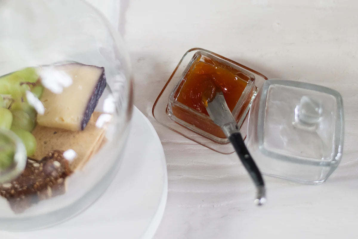 Square jar of jam with a knife next to a dessert with grapes and cheese on a white surface