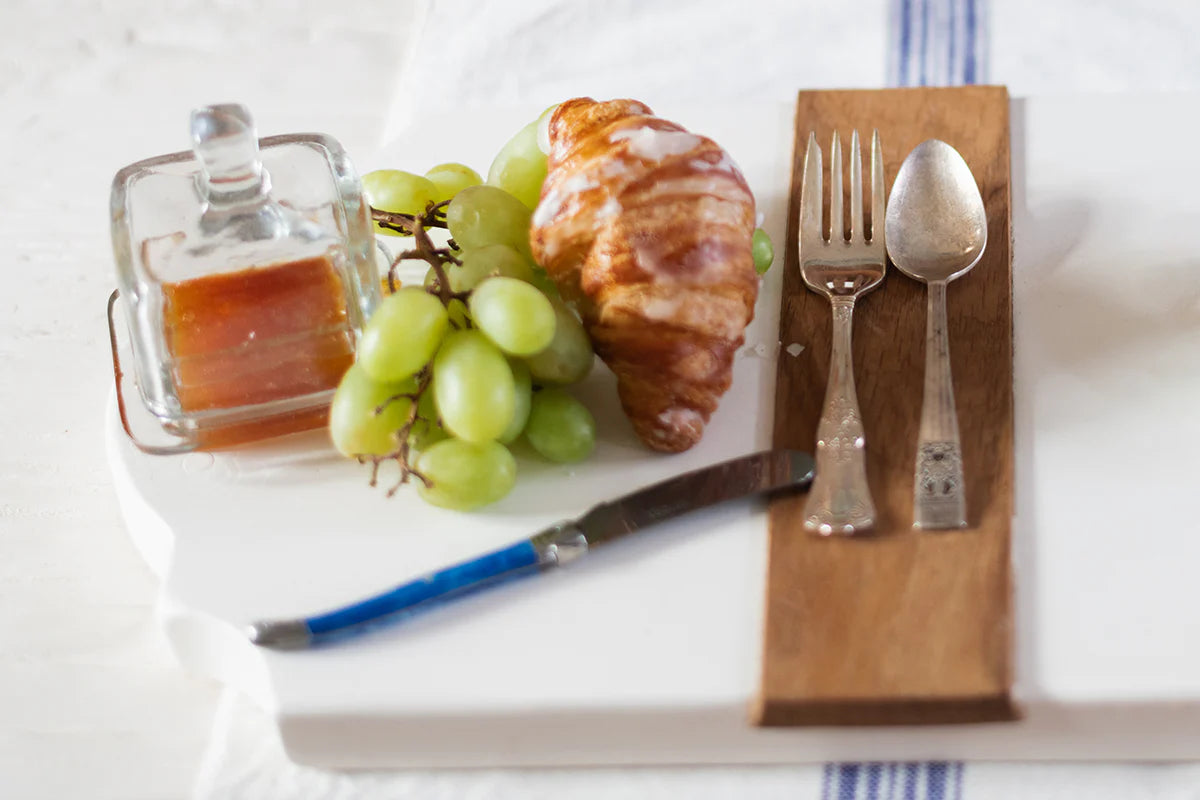 Breakfast setting with croissant, grapes, syrup, square jam jar, knife, and cutlery on a white surface.