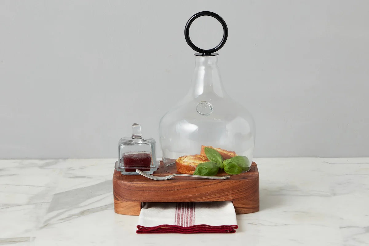 Glass cloche over bread with a square jam jar beside it on a with wooden serving board on a marble surface