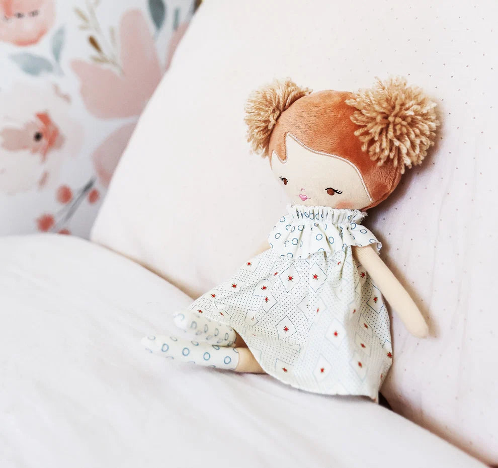 A plush doll with brown yarn hair in pigtails sits on a white bed. The doll is wearing a white dress with a blue and red geometric pattern.