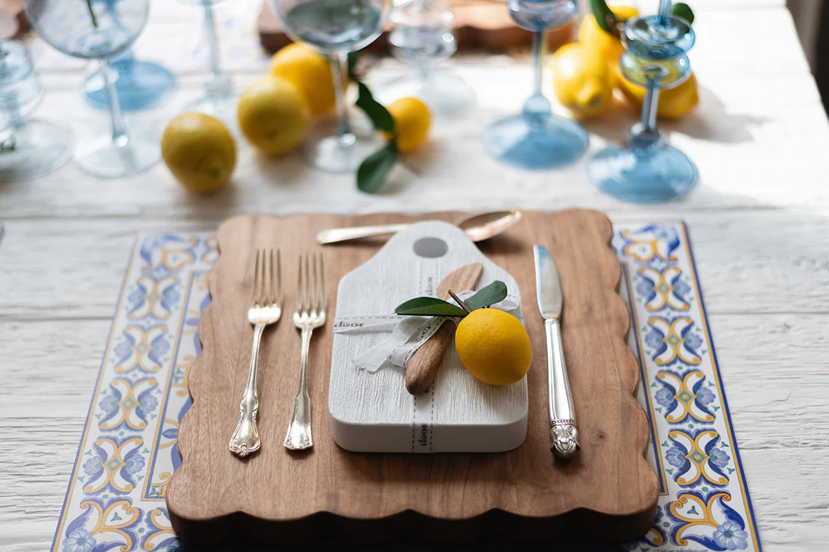 Table setting with white cheese serving board, utensils, and lemons on a decorative tablecloth.