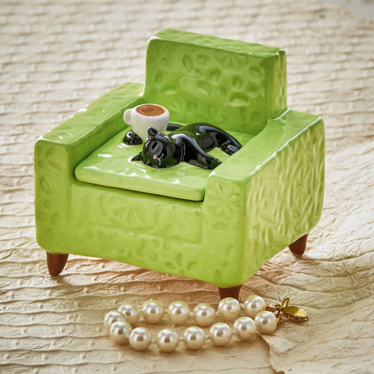 Green ceramic armchair with a black cat figurine and white cup, accompanied by a pearl necklace on a textured surface.