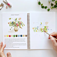 Person painting a floral design in an open art book with a poppy bouquet illustration.