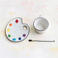 Paint palette shaped saucer with colorful paint swatches, a mug, and a brush shaped stirrer on a light background.