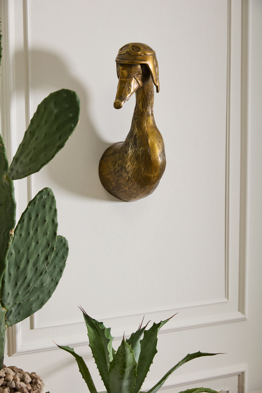 Decorative brass duck head on a white paneled wall with plants in the foreground