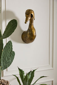 Decorative brass duck head on a white paneled wall with plants in the foreground