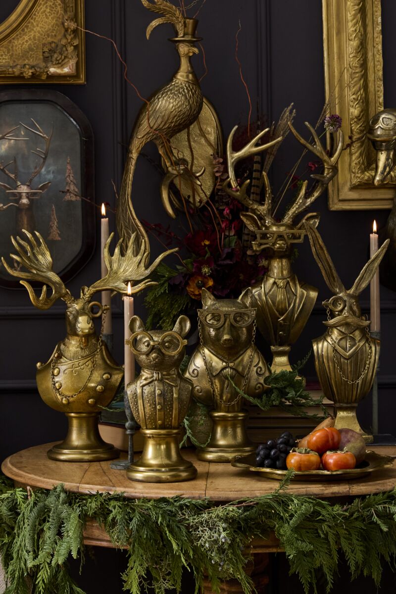 A collection of antique gold animal busts, all of them wearing various suits, glasses, and monocles on a wooden table with decorative greenery.