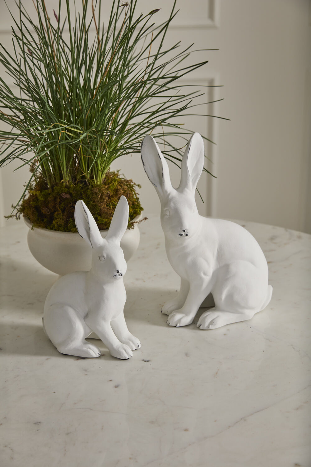 Two white rabbit sculptures on a light surface with a plant in the background.
