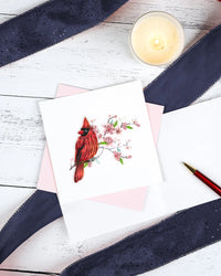 Card with a quilled red cardinal illustration on a white surface with navy ribbon and a lit candle.