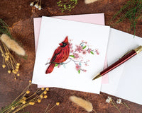 Card with a quilled red cardinal illustration on a rustic surface with a pen and flowers.