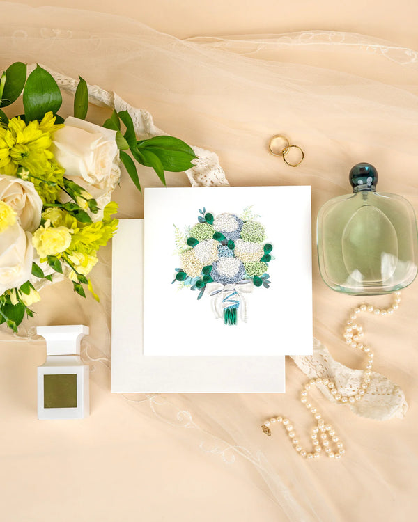 Floral arrangement, card with quilled hydrangea design, perfume bottle, and jewelry on a beige surface