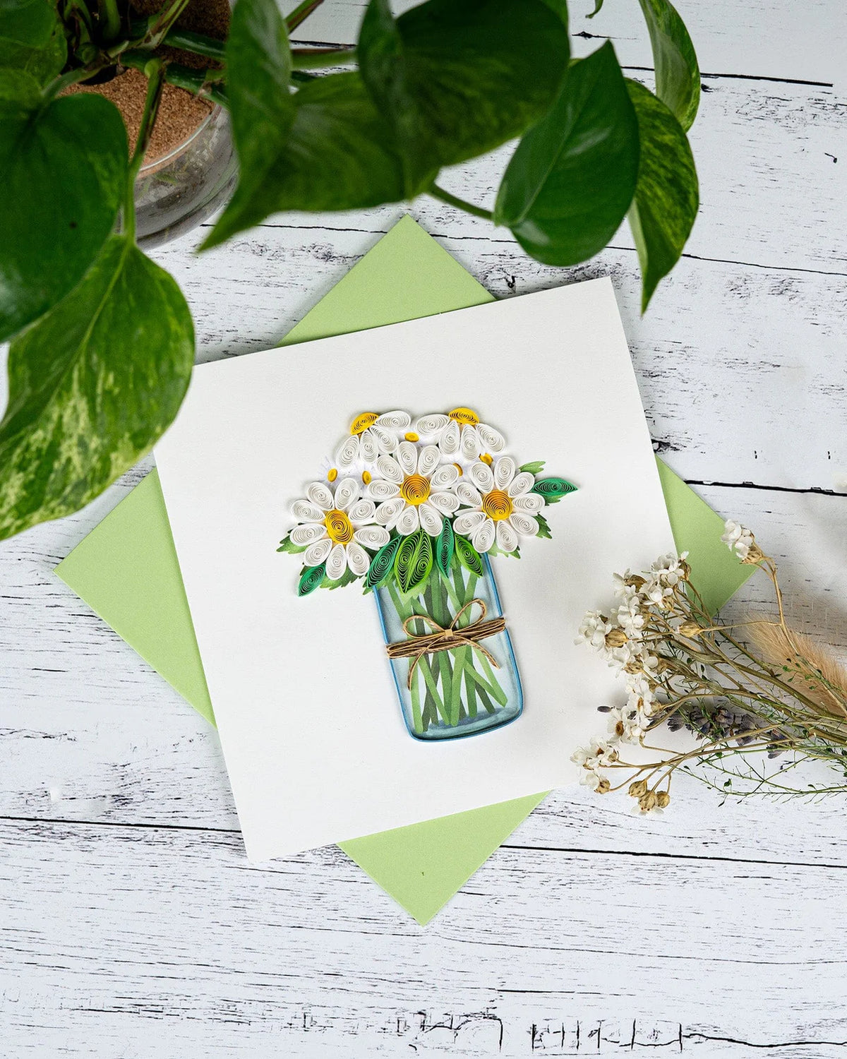 Card with quilled daisy design on a wooden surface with green leaves and flowers.