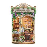 A decorative miniature diy book nook set labeled 'Impressionist Studio' shown against a plain white background. The scene includes a detailed storefront facade with shelves, flowers, plants, lamps, and a central arched window area.