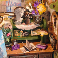 A vintage-style miniature display featuring a green shelf with a white lace doily, a silver floral vase of purple and yellow flowers, a small brown textured pot, and a sculpted bust. A wooden table below holds an open vintage book, a purple book, a small lamp, and other miniature decorative items.