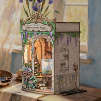 A decorative miniature art studio diorama set on a wooden table, featuring a detailed facade labeled 'Impressionist Studio' with a painted interior, floral accents, and a side panel with wisteria and wall decor. The scene is styled in a painterly room with blue walls, a window, and flowers in the background.