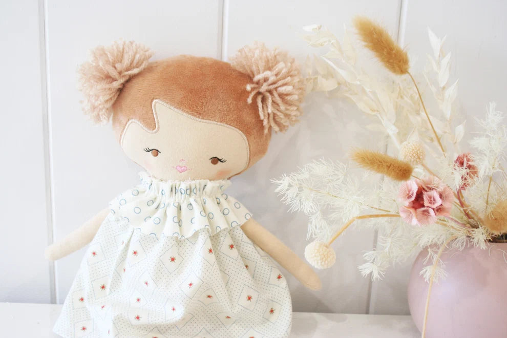 Stuffed doll with brown hair and a white dress next to dried flowers on a white background