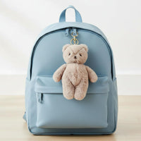A light blue nylon backpack with a front zippered pocket, featuring a small beige plush teddy bear keychain hanging from the zipper on a light wood floor against a white wall.
