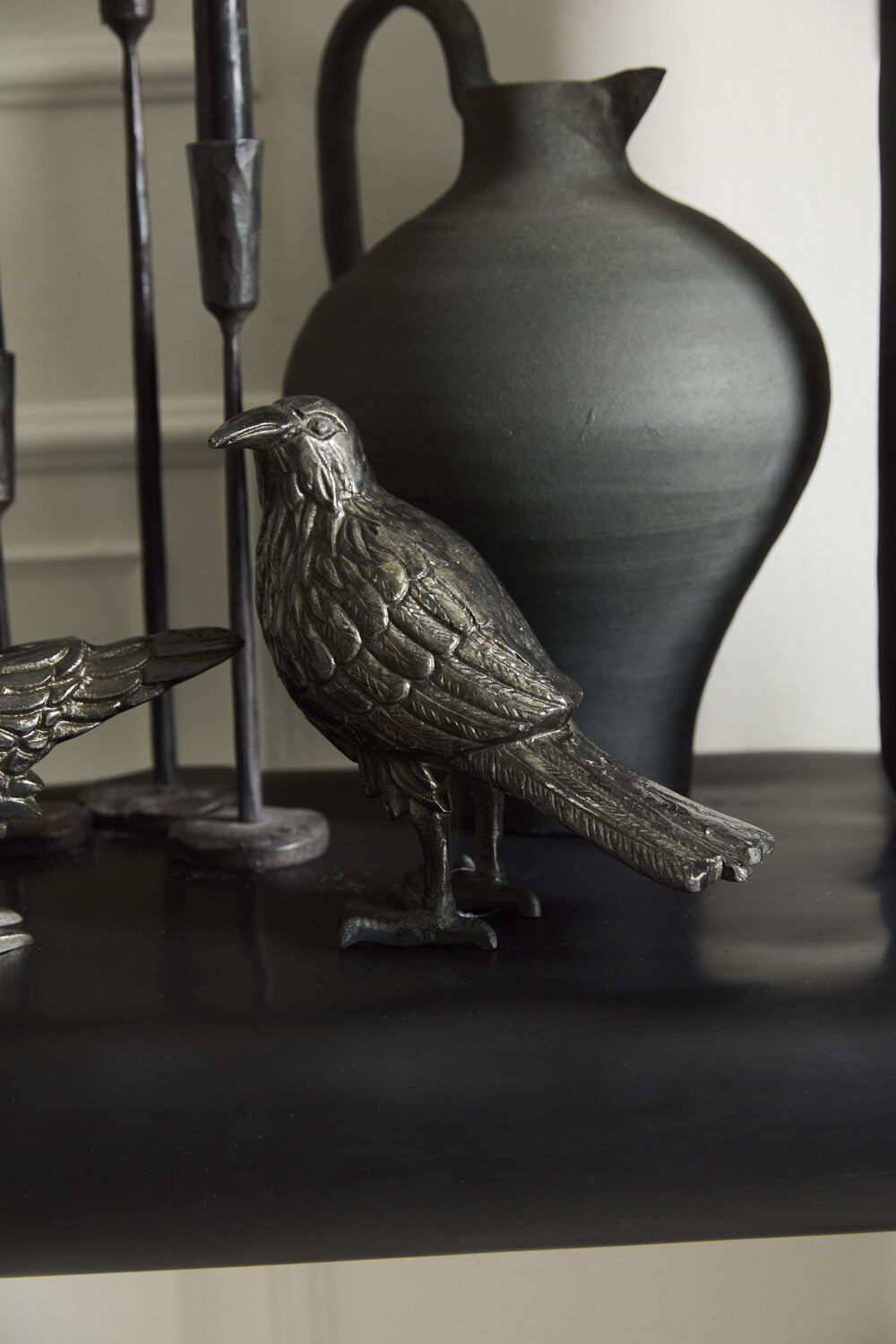 Decorative metal raven figure on a dark surface with a black vase in the background.