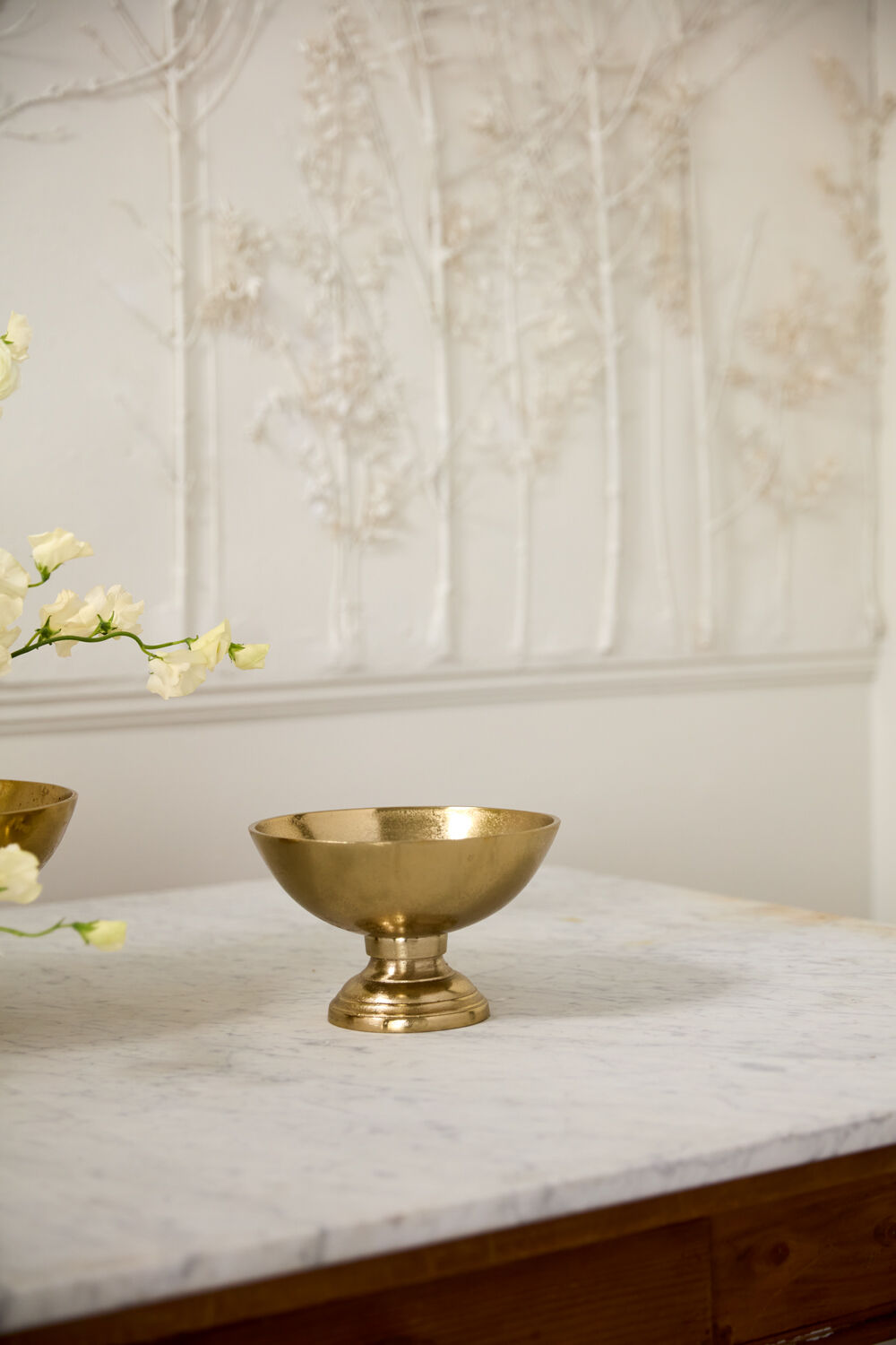 Gold bowl on a marble surface with floral decorations against a decorative wall.