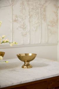 Gold bowl on a marble surface with floral decorations against a decorative wall.