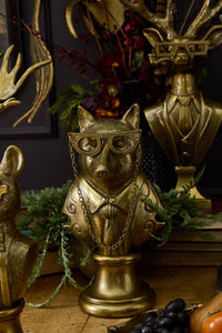 Gold animal head sculpture with glasses and chain, surrounded by greenery and other decorative items.