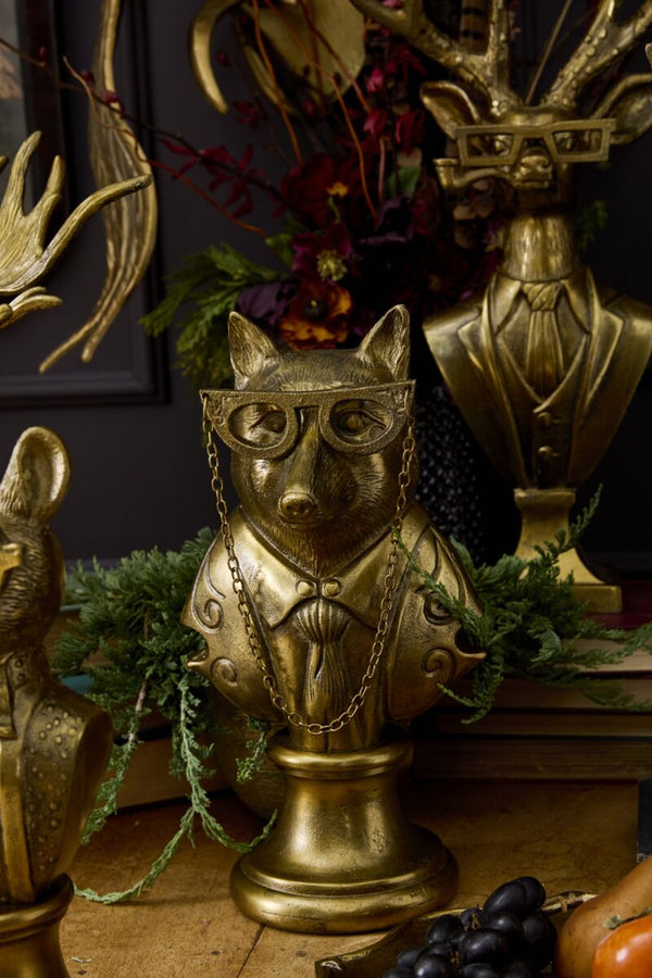 Gold animal head sculpture with glasses and chain, surrounded by greenery and other decorative items.