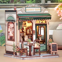 A miniature wooden dollhouse cafe model with a 'Sunny Cafe' facade, featuring a green awning, detailed interior, outdoor seating, and potted plants, displayed on a tabletop.