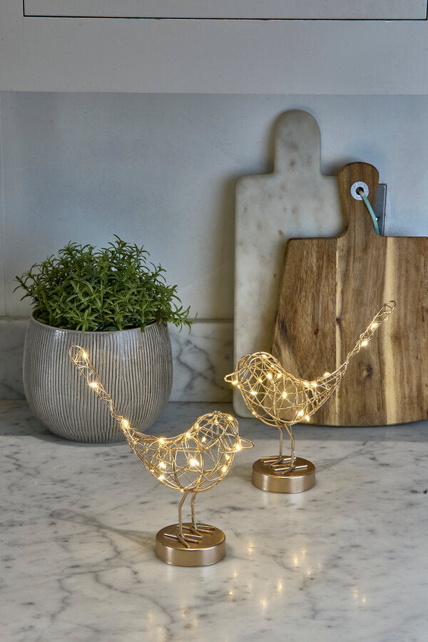 Decorative gold bird sculptures with LED lights on a marble surface, with a plant and cutting board in the background.