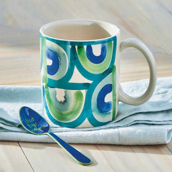 Mug with green and blue circular pattern on a light wooden surface with a spoon.